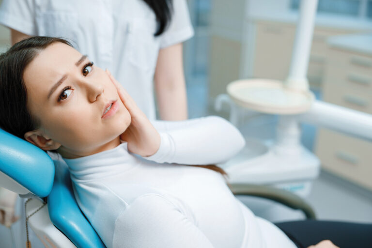 Patient in dental chair. Beautiful young woman having dental treatment at dentist's office. Girl touch her cheek suffering from painful toothache.