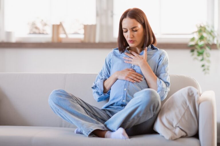 Pregnant lady feeling bad having discomfort and touching her tummy and chest, coping with abdominal spasms and asthmatic issues in living room at home. Complexities of expectant motherhood
