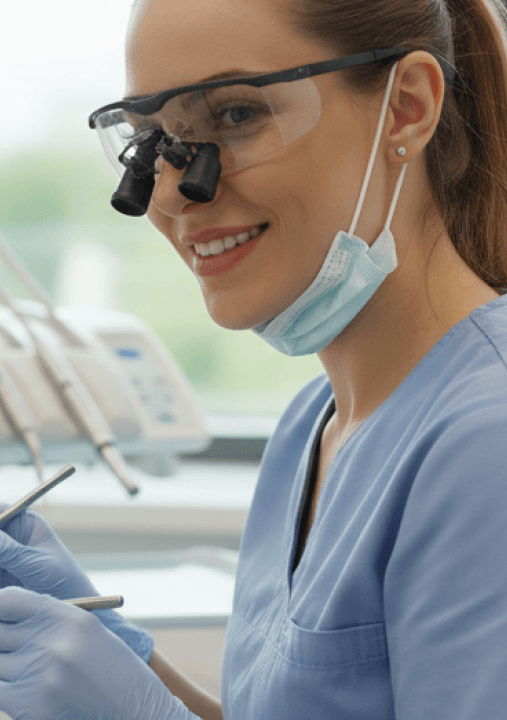 A dentist wearing loupes and a surgical mask preparing to examine a patient, illustrating comprehensive oral health care